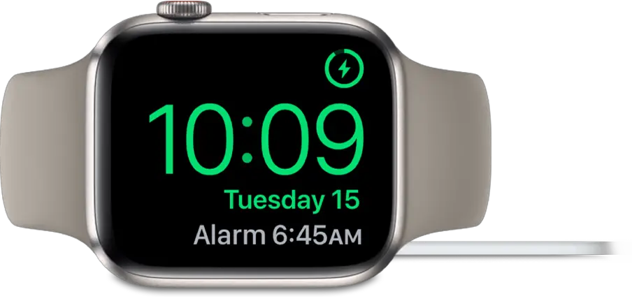 An Apple Watch placed on its side and connected to the charger, with the screen showing the charging symbol in the top-right corner, the current time below that, and the time of the next alarm.